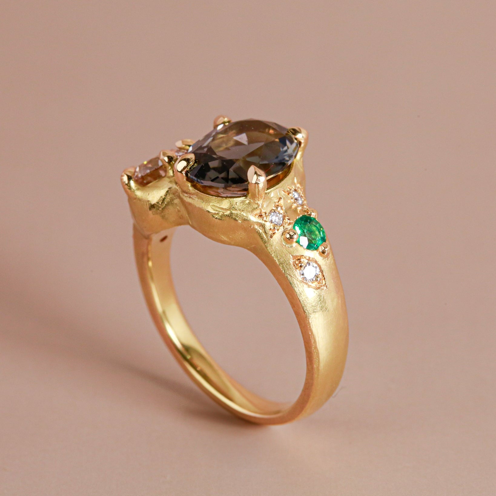 Gold ring with a central dark gemstone and smaller green and white gemstones on a beige background