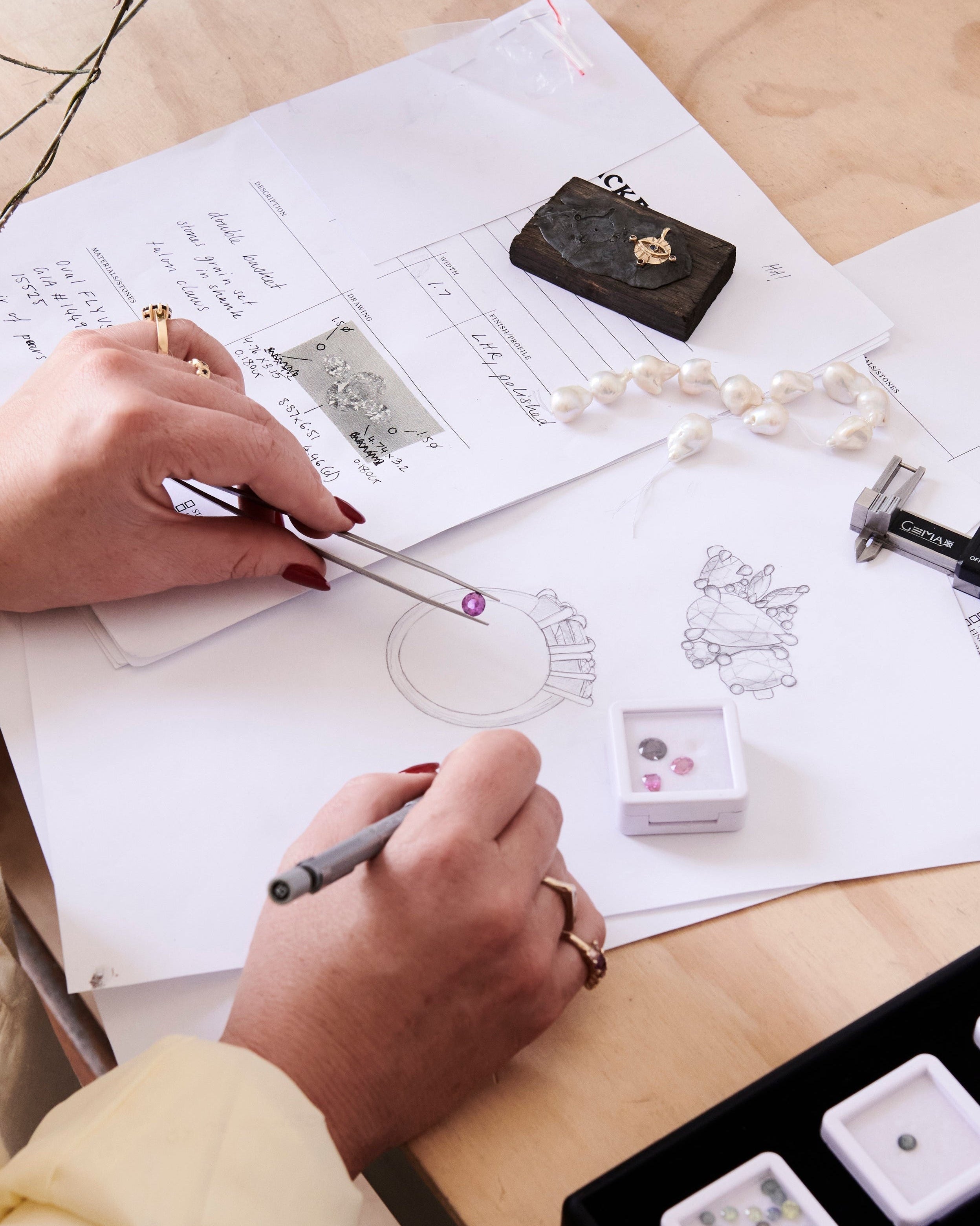 Person working on jewelry design with sketches and materials on a table.