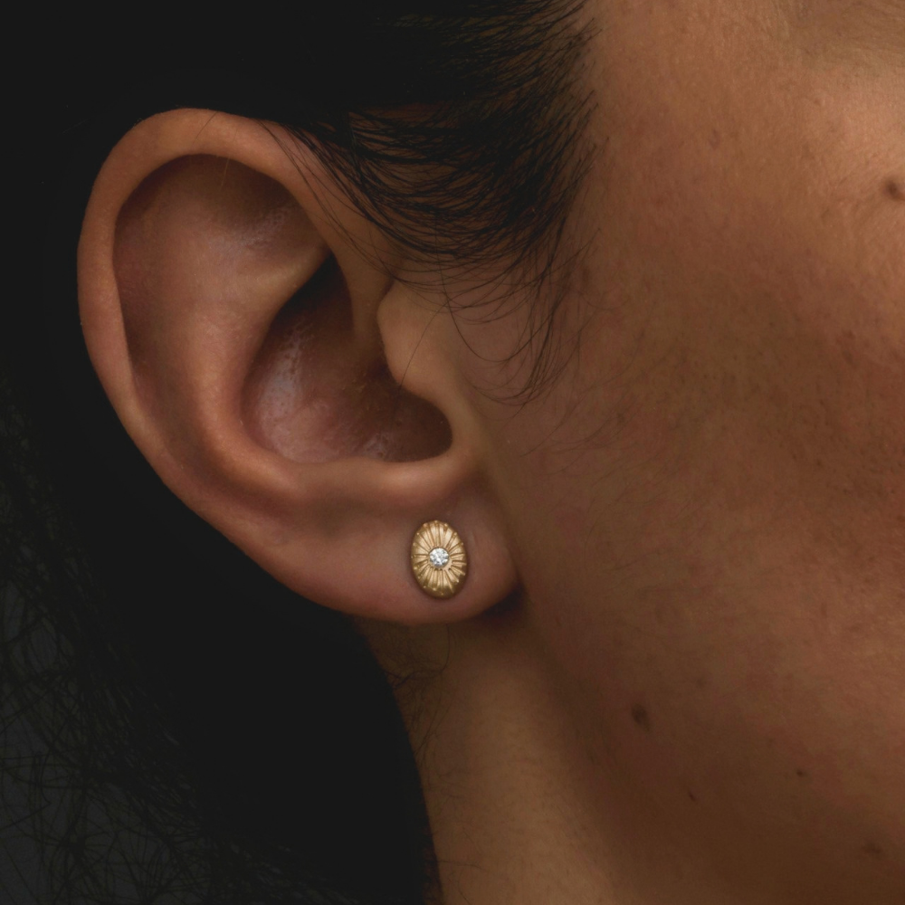 A close-up of a person's ear wearing a small gold post earring with a textured sunburst design and a tiny diamond centre stone.