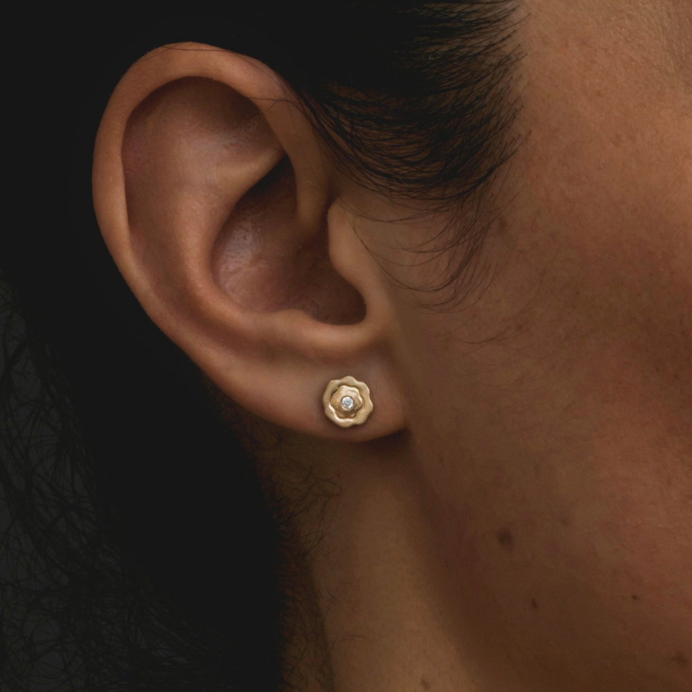 A close-up of a person's ear wearing a small gold stud earring with a clear central gemstone, set against a dark background.
