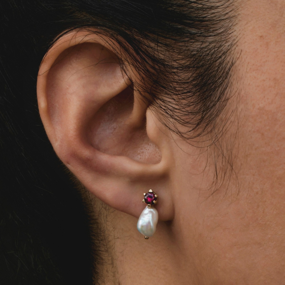 A close-up of a person's ear wearing a small drop earring with a white baroque pearl and a small red garnet in a gold setting.