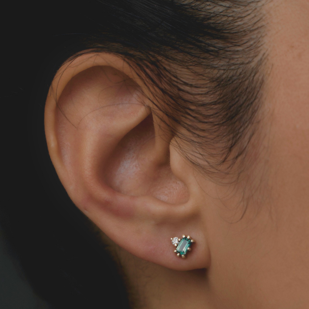 Close-up of a gold stud earring worn on the ear, featuring a rectangular green gemstone with a small white gemstone accent.