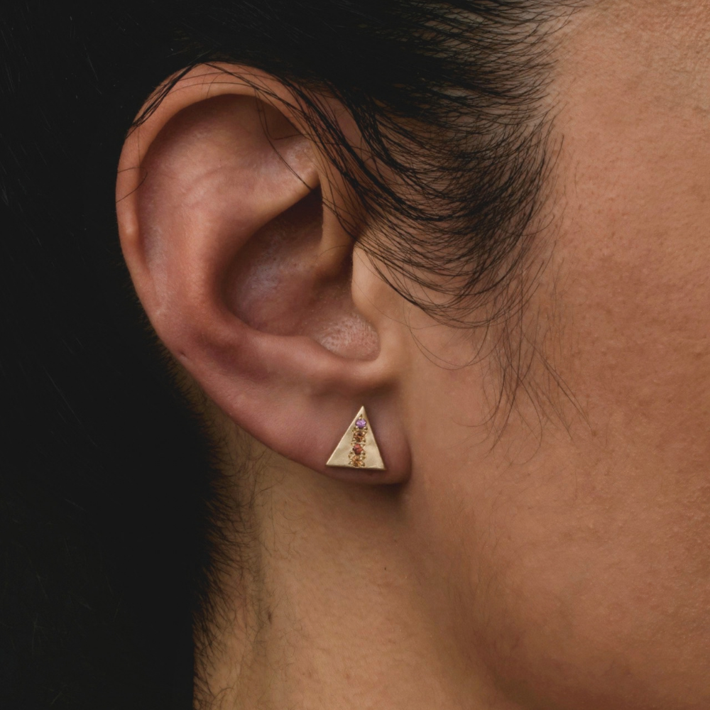 A close-up of a person's ear wearing a small triangular gold stud earring with a central vertical arrangement of purple and red sapphires.