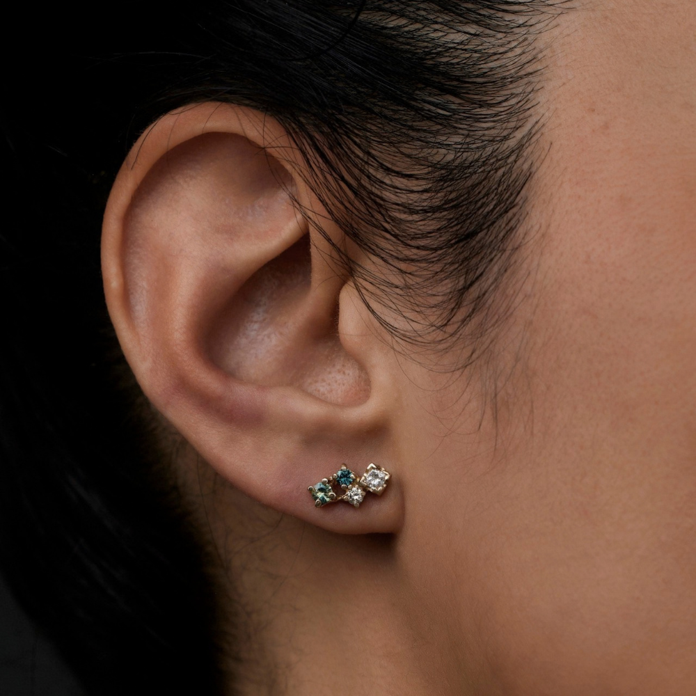A close-up of a person's ear wearing a small cluster stud earring with green sapphires and white diamonds.