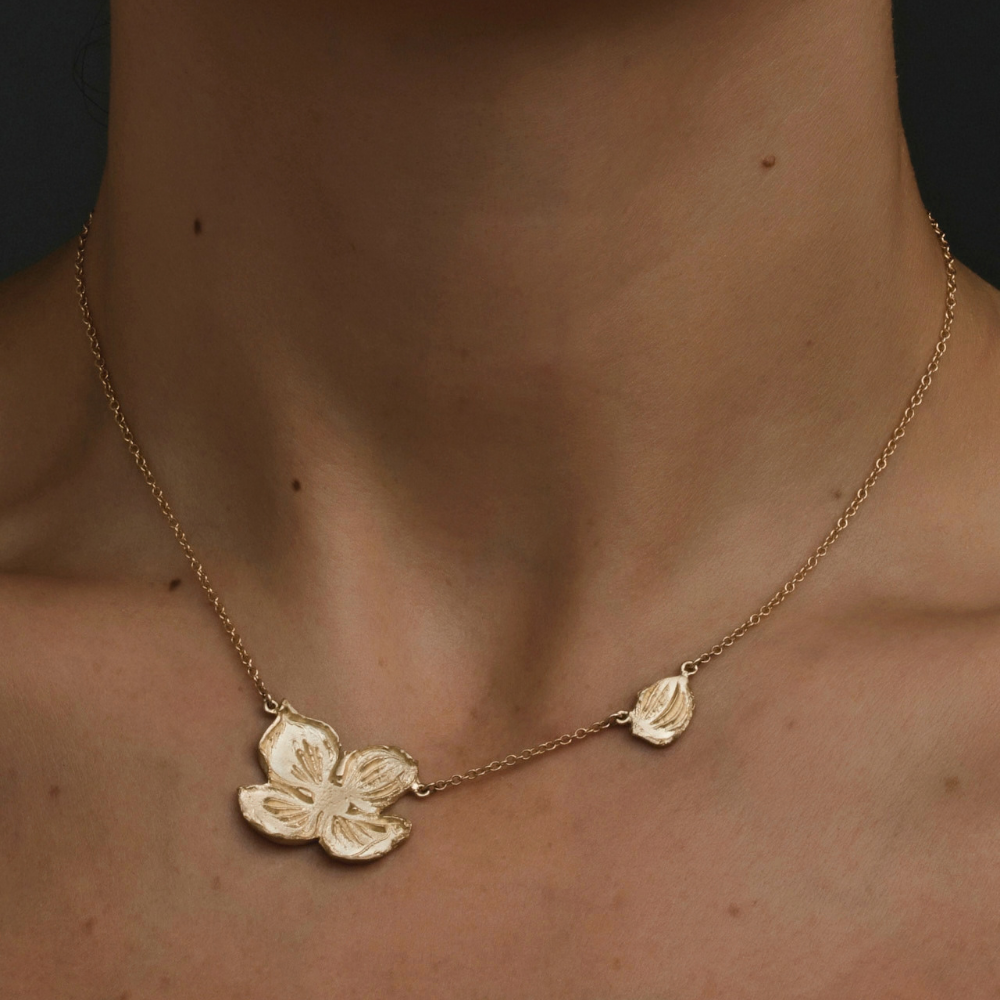A close-up of a person's neck wearing a delicate gold chain necklace with a large flower-shaped pendant and a smaller leaf-shaped pendant.