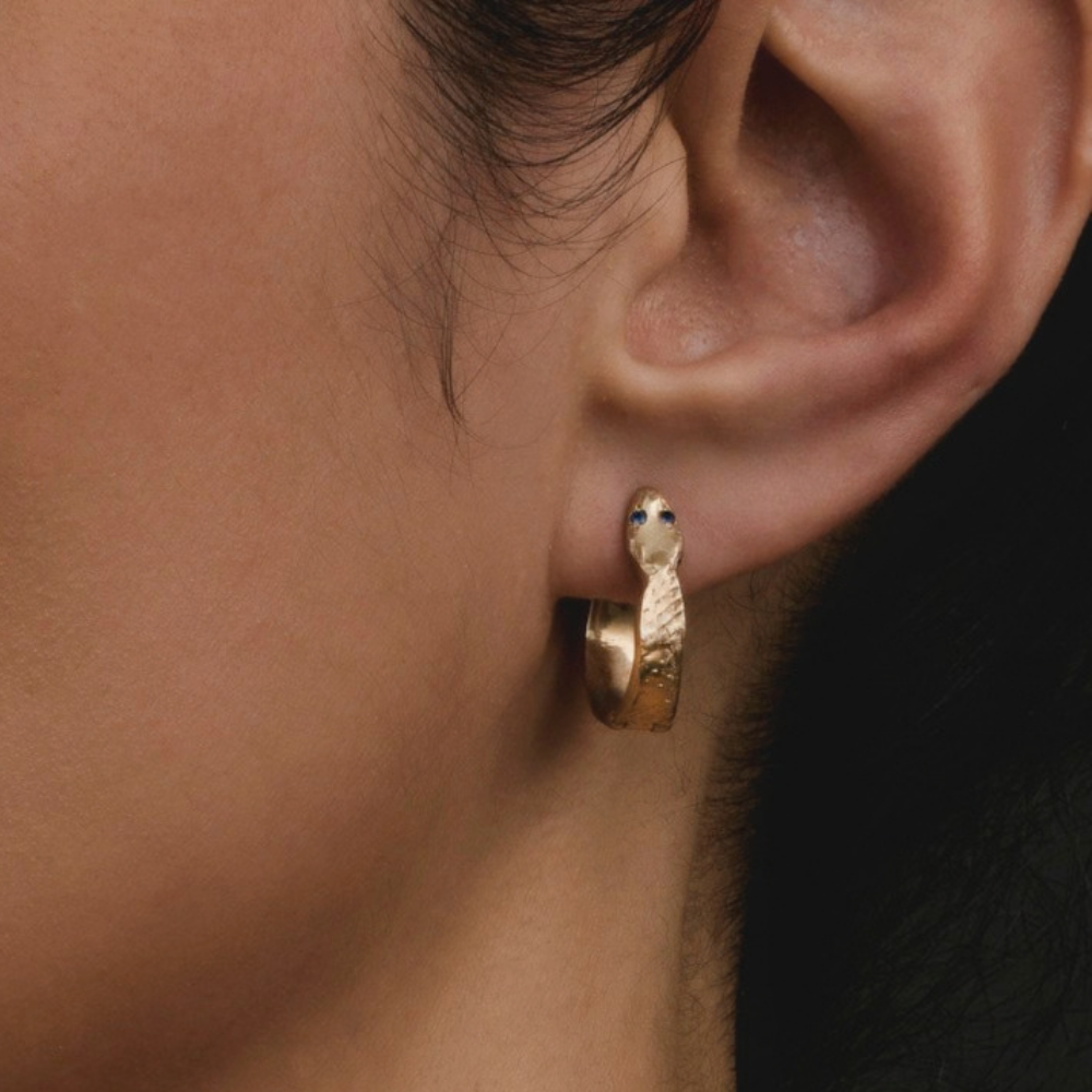 a close up of a person's ear wearing a medium sized gold hoop huggie earring shaped like a snake with blue gemstone eyes.